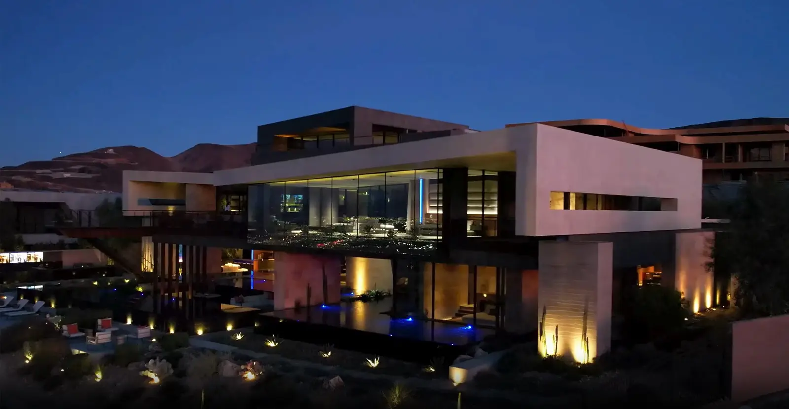 A modern, multi-level house illuminated at night takes center stage in this architectural slideshow. With its large glass windows and geometric design, it is surrounded by landscape lighting that enhances the warm ambiance against the dark blue evening sky.