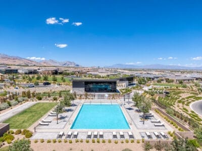 An outdoor swimming pool surrounded by a patio with lounge chairs and a neatly landscaped area, featuring a modern building in the background. The backdrop includes mountains and a clear blue sky, part of the stunning Mesa Ridge development. Multiple buildings and green spaces are visible in the distance.