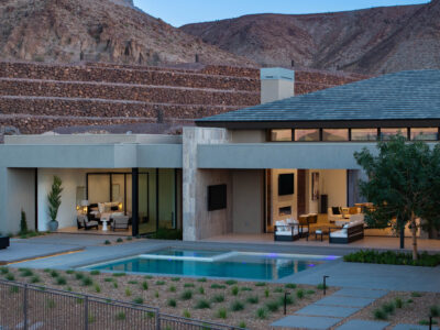 Modern house on Rockmount Court with an extensive patio and pool, set against a backdrop of terraced hills. The house features large glass windows, allowing a clear view of the contemporary interior with plush seating and a minimalist aesthetic. The outdoor area is landscaped with greenery.