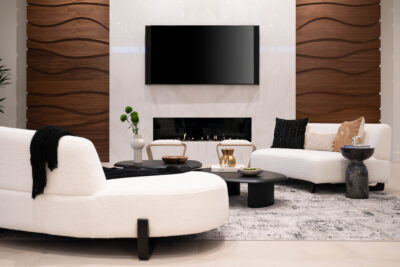 A modern living room in Ascaya with white curved sofas, a sleek coffee table, and a fireplace beneath a large flat-screen TV. The wall features wood paneling with wavy patterns, and there are decorative items such as vases, cushions, and a rug adding texture to the space.