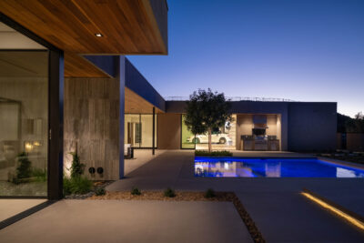 A modern home at 6 Rockmount Court is illuminated at dusk, showcasing its sleek architecture. The backyard features a swimming pool with blue lighting, a tree, and an outdoor kitchen area. Large glass doors open to the exterior space, highlighting the contemporary Ascaya design.