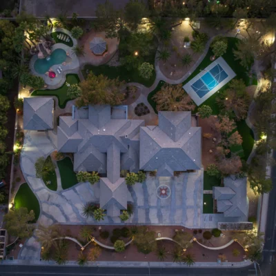 Aerial view of a large residential property at dusk, featuring a multi-story house with a gray roof, a swimming pool with a slide, and a basketball court. The lush gardens with trees and pathways add charm, complemented by several smaller structures. The entire scene benefits from strategic outdoor lighting that seems almost auto-draft in its perfection.