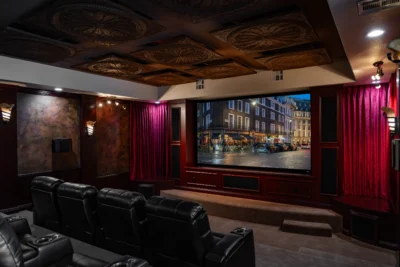 A home theater features a large screen displaying a cityscape at night, surrounded by red curtains. The room, equipped with leather recliners in rows and ornate ceiling tiles, also has wall sconces and decorative finishes. With dim lighting creating a cozy atmosphere, it feels like an auto draft of comfort and style.