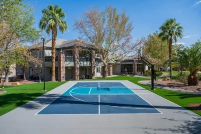 A modern outdoor basketball court with a blue and gray surface is situated in front of a large house, designed with an auto draft layout. The area is surrounded by trees, palm trees, and a well-maintained lawn under a clear, blue sky.