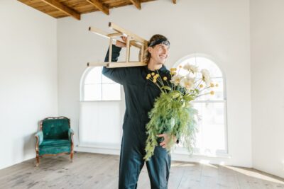 A person wearing a black jumpsuit and a bandana smiles while holding a wooden stool over their shoulder and a large bouquet of yellow and white flowers in their other arm. They stand in a newly developed, brightly lit room with arched windows and a green armchair in the corner.