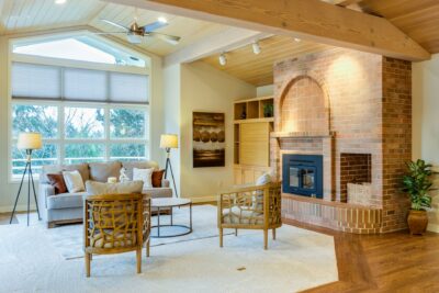 A cozy living room with a brick fireplace, a gray couch adorned with pillows, two wicker armchairs, and two floor lamps. Large windows let in natural light. The space includes wooden beams and a vaulted ceiling, emulating best practices found in luxury communities—a small plant sits in the corner near the fireplace.