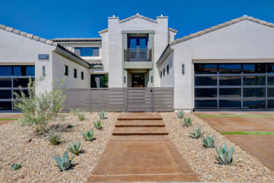 This modern white house, reminiscent of Luxury Homes in Red Rock Country Club, boasts a central entrance flanked by two large garages. The front features desert landscaping with gravel and succulents under a clear Las Vegas sky.