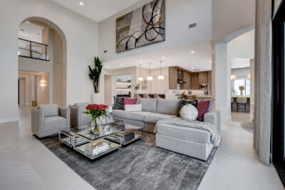 Spacious modern living room in a luxury home at Red Rock Country Club, showcasing a large gray sectional sofa, glass coffee table, and abstract art. The well-lit area boasts an open-plan view of the kitchen and dining area. Neutral tones with red floral accents add warmth.