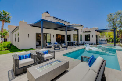 A modern white luxury home in Las Vegas boasts large windows and an outdoor patio with a swimming pool, lounge chairs, and a dining area. Adorned with gray furnishings and blue cushions, the serene setting is reminiscent of the opulence found at Red Rock Country Club.