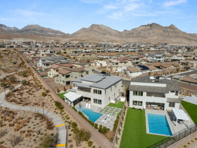 Aerial view of a guard-gated Summerlin neighborhood with modern houses, two homes featuring backyard swimming pools, green lawns, and a walking path next to desert landscaping, with mountains in the background under a blue sky.