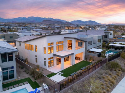 Modern two-story house with large windows and patio, warmly lit at dusk in guard-gated Summerlin. Landscaped yards frame the home, neighboring modern houses, and mountain views under a colorful sky.