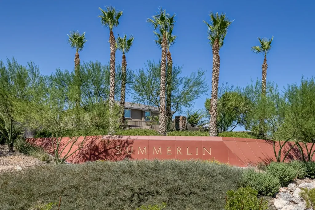 A landscaped Summerlin entrance with palm trees and desert plants features a large red sign that reads "SUMMERLIN" beneath a clear blue sky.