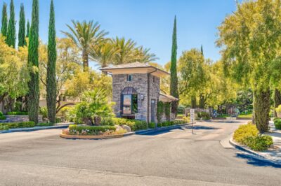 A stone security gatehouse marks the entrance to this guard-gated Summerlin community, surrounded by tall trees, manicured landscaping, and palm trees beneath a clear blue sky.