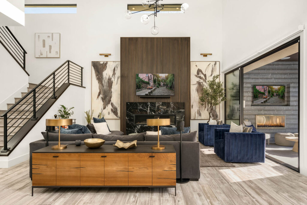Modern living room in The Ridges with a black marble fireplace, wooden accent wall, blue velvet chairs, and neutral decor. Large glass doors open to an outdoor patio with a fireplace. Stairs with black railings lead to an upper floor.