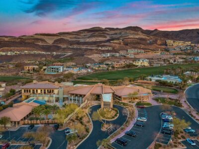Aerial view of a large desert residential community at sunset, with modern houses, a green golf course, winding roads, parked cars, and mountains in the background.