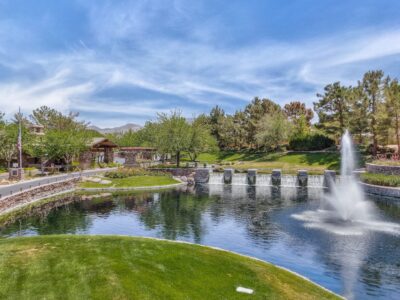 A landscaped park with a pond, a row of small waterfalls, a large fountain, green lawns, trees, and distant mountains under a blue sky with wispy clouds.