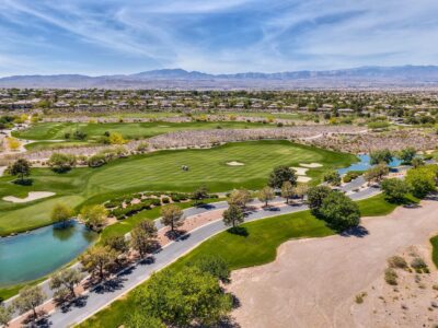 Aerial view of a lush green golf course with sand bunkers, small ponds, trees, and a golf cart. Residential homes and mountains are visible in the background under a blue sky.