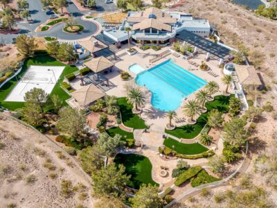 Aerial view of a large recreation center with a rectangular swimming pool, lounge chairs, cabanas, shaded areas, landscaped gardens, palm trees, and nearby parking, surrounded by desert terrain.