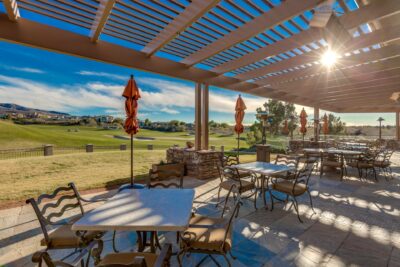 Outdoor patio with tables and chairs under a pergola, overlooking a sunny golf course with mountains and houses in the distance; orange umbrellas are closed, and sunlight filters through the slats.
