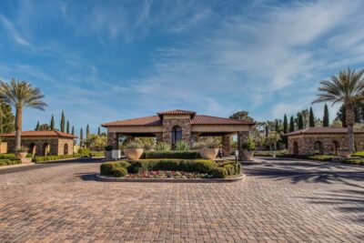 A Mediterranean-style building with a red-tiled roof sits at the center of a circular driveway, surrounded by manicured bushes, palm trees, large planters, and vibrant flower beds under a blue sky.