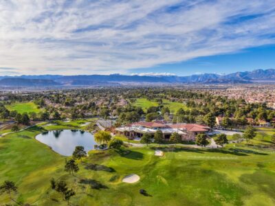 Aerial view of a golf course with a large clubhouse, pond, and manicured greens, surrounded by trees and residential houses. Mountains and a blue sky with scattered clouds are visible in the background.