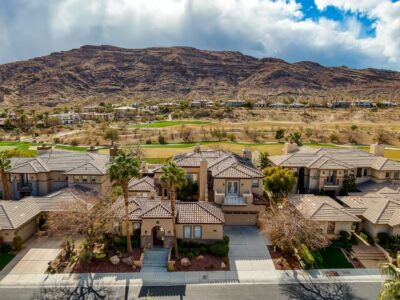 Aerial view of a row of modern, tan-colored houses with tile roofs, palm trees, and manicured lawns; a golf course and rocky hills are visible in the background under a partly cloudy sky.