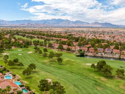 Aerial view of a golf course with green fairways, sand bunkers, and trees, bordered by houses and a canal, with mountains and a cityscape in the background under a partly cloudy sky.